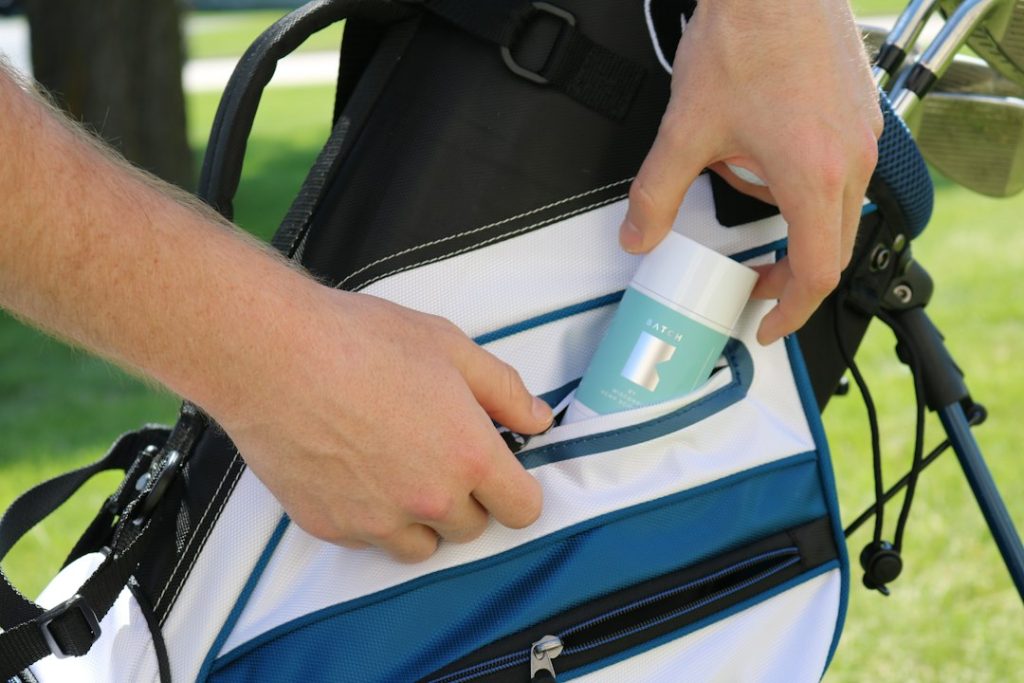 Hands pulling CBD balm out of pocket of golf bag on golf course.